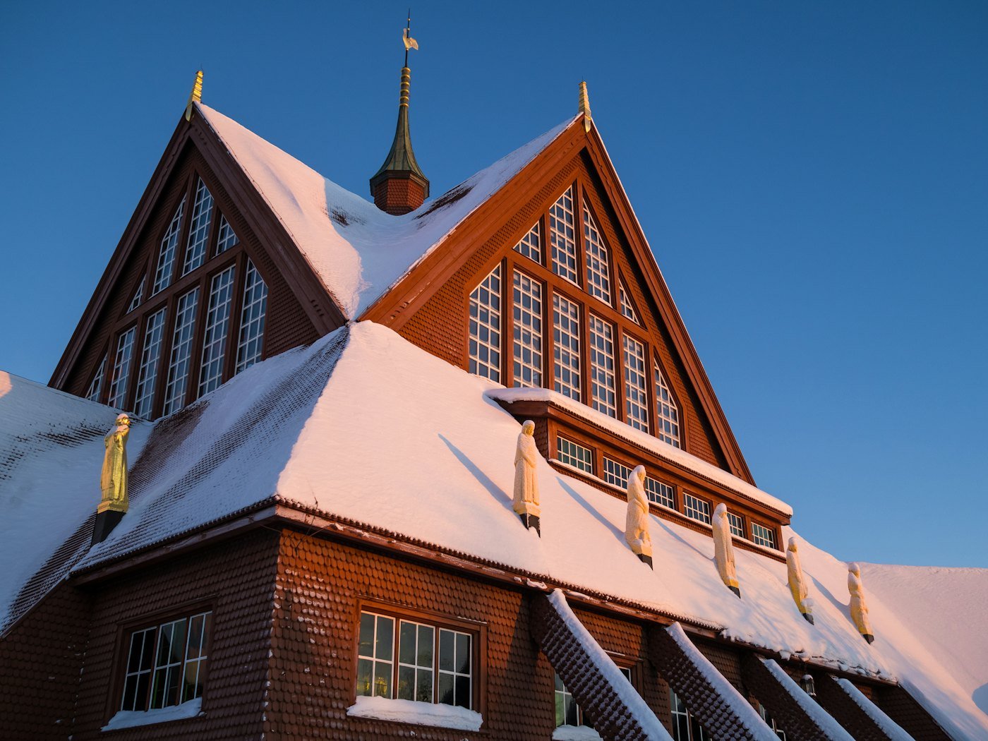 Steep wooden gables of Kiruna Church under snow, gilded statues along the roofline in low sun