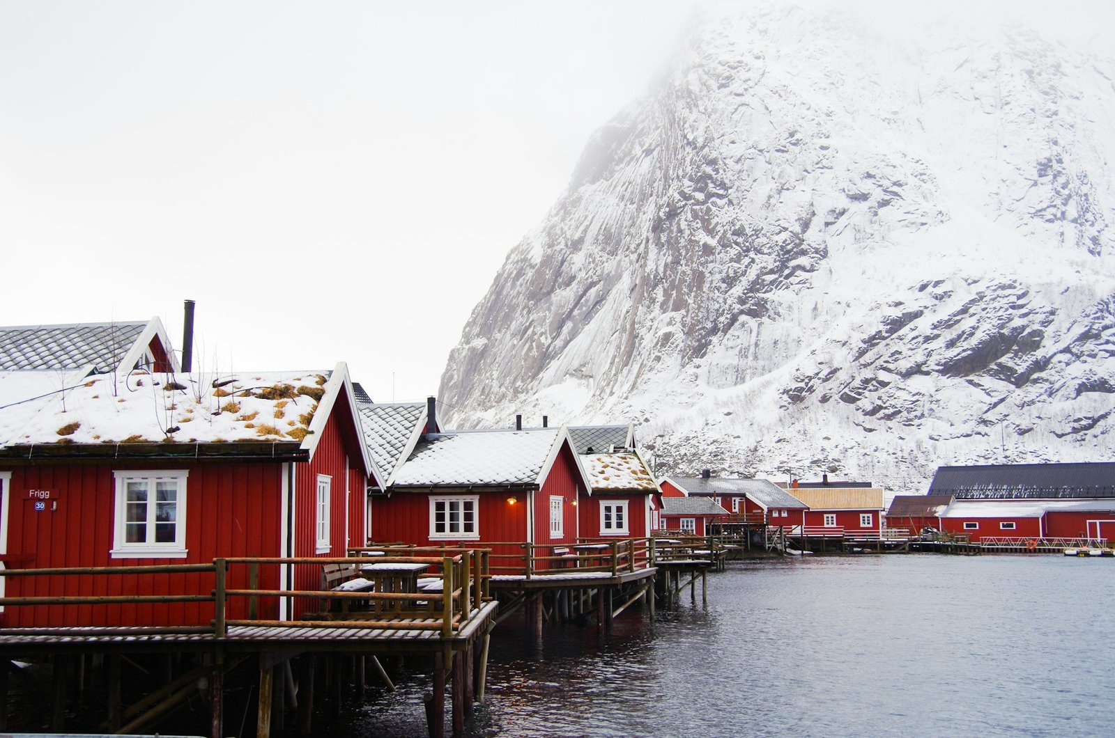 Reine village, red rorbuer cabins against steep snow-covered mountains, water in foreground
