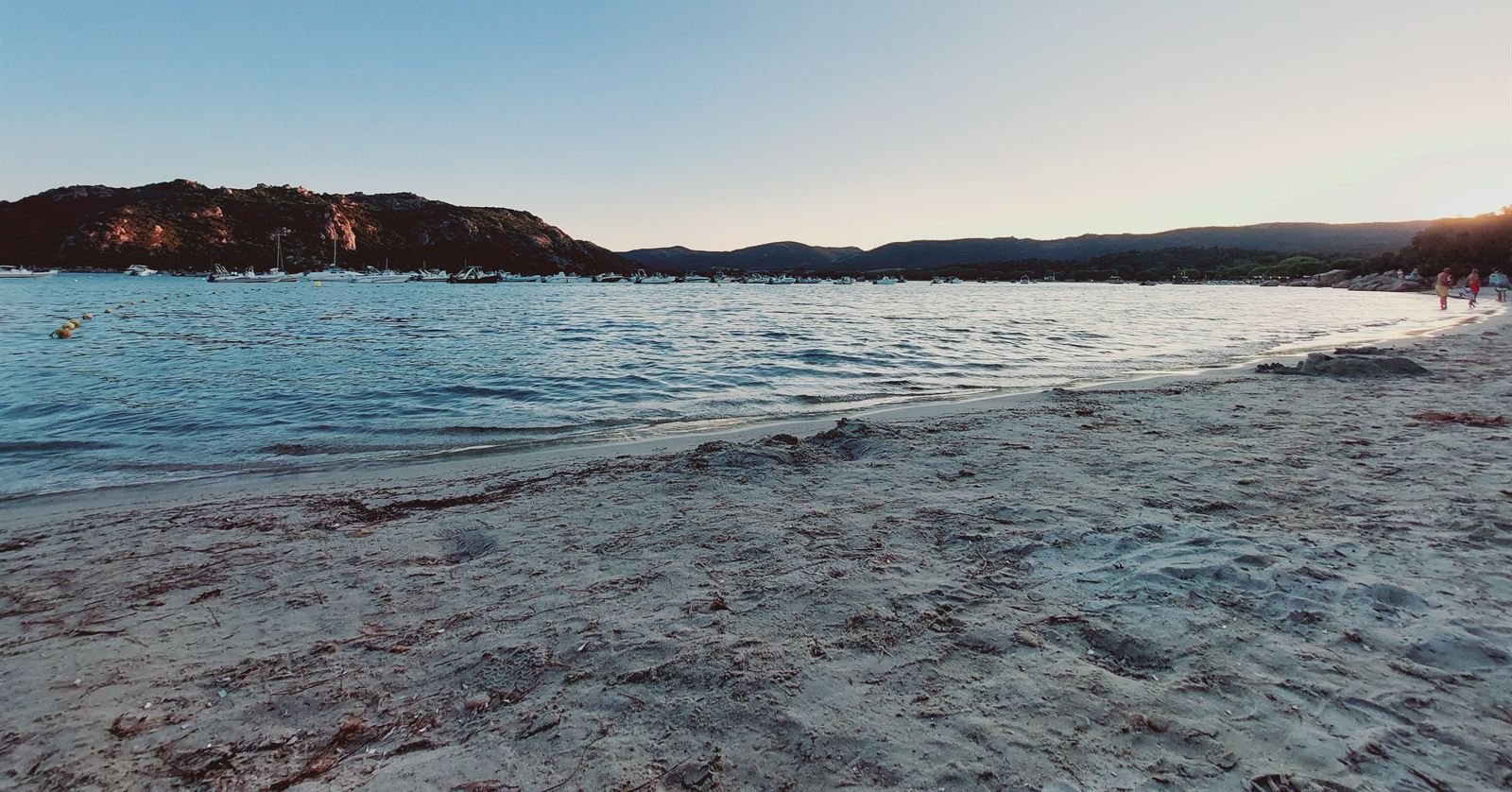 A quiet beach at dusk, small boats moored off a rocky headland